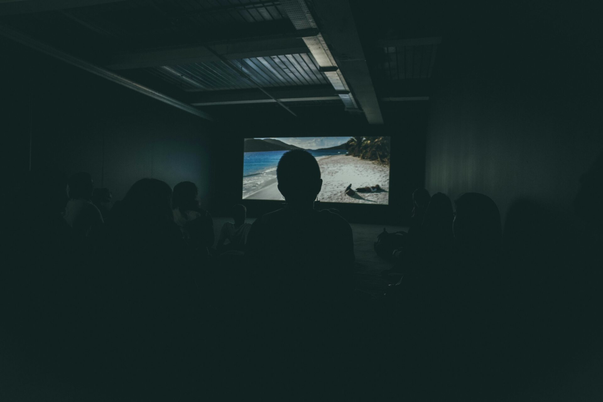 An image of a dark room with a tv screen and people sat in front watching it.